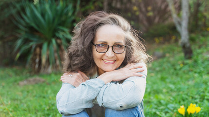 Obraz premium Portrait of a beautiful senior woman 60 years old in glasses with a charming smile looking at the camera while sitting on the grass in the garden
