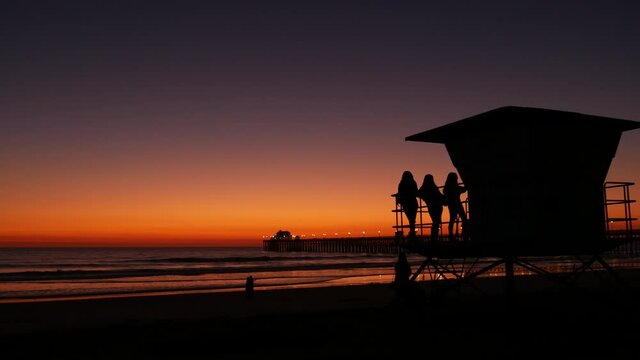 Young teen girls silhouettes near lifeguard tower, friends on pacific ocean beach, sunset dusk in Oceanside, California USA. Unrecognizable teenagers, people and twilight gradient purple violet sky.