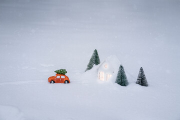 Orange toy car carrying Christmas tree on roof in the snow near toy house among trees
