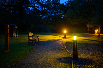 A park at night. A stony path cuts across a lawn and is illuminated by electric lights. The lamps cast a yellow light and long shadows. Focus on the bench with shallow depth of field.