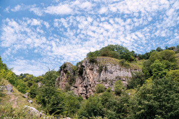 White feathery clouds in light blue sky above a rocky cliff covered with trees and bushes