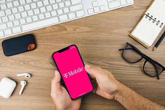 Male hand holding a smartphone with T-Mobile app on the screen with a computer keyboard. Wooden Background. Mobile communications company.Rio de Janeiro, RJ, Brazil. October 2021.