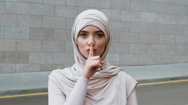 Outdoor portrait of young confident muslim woman wearing hijab putting finger on lips, showing shh gesture