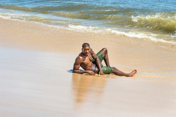 A well built young  black guy, half naked,  is  lying on the beach and playing with waves of water..