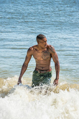 A well built young  black guy, half naked,  is standing in water on the beach and looking away..