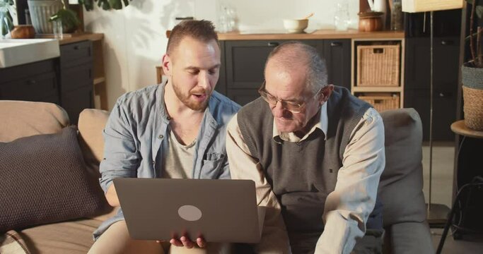 Happy bearded 30s young adult man helping senior retired 70s dad use laptop and internet on couch at home living room.