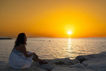 A woman in a white dress sits on the pier and admires the sunset on the sea