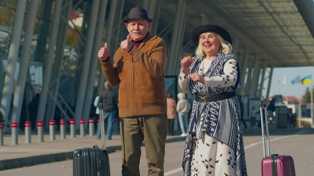 Elderly old husband and wife retirees tourists dancing disco fooling around having fun expressive gesticulating hands near airport terminal. Lovely mature couple grandmother grandfather celebrating