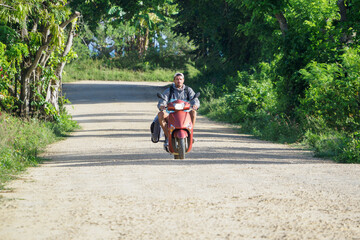 Man riding motor scooter with surfboard along dirt track next to Ocean.