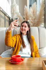 Happy woman in yellow cardigan and long hair is taking selfie on mobile phone in cafe.