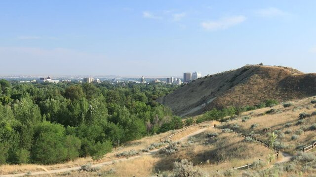 A view of downtown Boise, Idaho as seen from Camel’s Back Park on a sunny, summer day. A  network of sandy trails is seen crisscrossing foothill slopes covered in dry grasses.