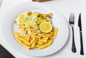 Roasted sepia served with fried potatoes, piquant green sauce and lemon on white plate