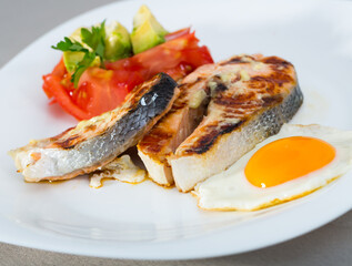 Delicious roasted salmon and fried egg served with fresh vegetables and greens on white plate..