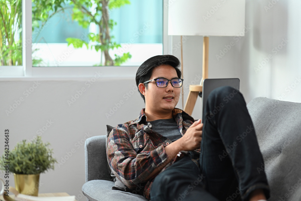 Poster smiling asian man laying on sofa and browsing internet with digital tablet.