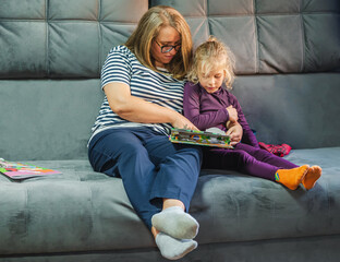 A woman in glasses is a mother reading a picture book to her child girl, sitting at home on a gray...