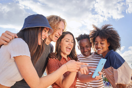 Group Of Young Mixed Race People Looking At A Cell Phone While Pointing At Something Funny On The Screen. Concept Of Technology, Connection, App, Online, Social Media.