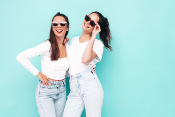 Two young beautiful smiling brunette hipster female in trendy summer clothes. Sexy carefree women posing near blue wall. Positive models having fun. Cheerful and happy in sunglasses