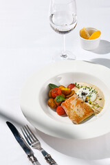 Fish steak with fillet tuna, warm tomato, baked potato and vitello tonnato sauce. Fish in white plate. Tuna sauce and vegetables. Contemporary seafood restaurant menu, summer day with sunlight shadow.