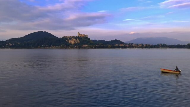 Small fishing boat with fisherman rowing in calm lake waters of Maggiore lake in Italy with Angera castle in background. Zoom out