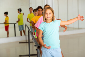 Cute cheerful preteen girl working near ballet barre during group choreography class.