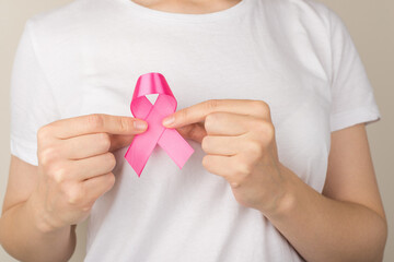 Close up photo of girl in white t-shirt holding pink ribbon symbol of breast cancer awareness on isolated grey background