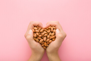 Heap of brown almond nuts in heart shape created from young adult woman hands on light pink table background. Pastel color. Closeup. Point of view shot. Top down view.
