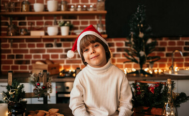 Obraz na płótnie Canvas portrait of a little boy in a white sweater and a red Christmas hat sitting on a table in the kitchen with