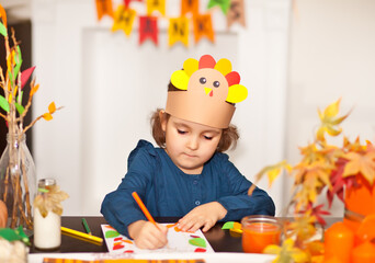 Little cute girl in paper turkey hat writing I am thankful for. Celebrating Thanksgiving day. Diy...