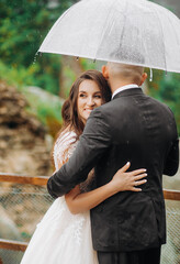 Cute bride and groom hugging in the mountains under a transparent umbrella. Raindrops. Wedding photo shoot on a background of rocks.