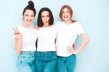 Obraz premium Three young beautiful smiling hipster female in trendy same summer white t-shirt and jeans clothes. Sexy carefree women posing near light blue wall in studio. Cheerful and positive models having fun