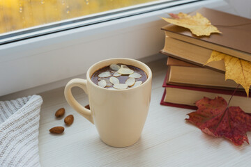 Fototapeta na wymiar Cup of hot chocolate with almond and books on the windowsill. Window in a background. Fall time. Autumn composition. Home comfort.