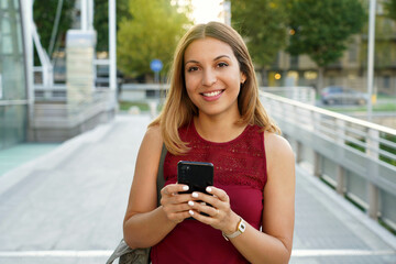 Smiling young woman is walking in the street with phone. She is holding gadget in hand and looking at camera. - obrazy, fototapety, plakaty