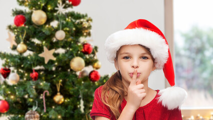 winter holidays and children concept - happy girl child in santa helper hat making shh gesture over...