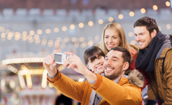 winter holiday, technology and leisure concept - happy friends taking selfie with digital camera over christmas market or amusement park background