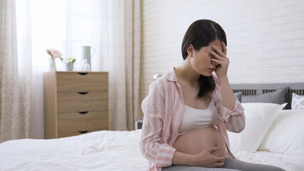 portrait Taiwanese woman sitting alone and covering her face in despair is suffering depression during pregnancy on bed at home.