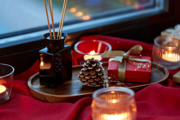 holidays, decoration and celebration concept - christmas gift, candles and aroma reed diffuser on red tablecloth on window sill at home