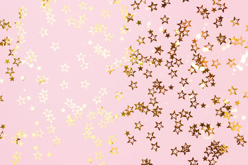 Metallic gold colored glittering stars confetti scattered on a pink pastel background.