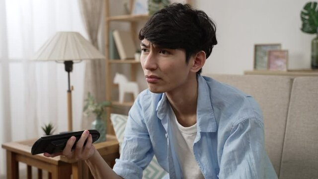 bored asian male turning off tv with remote control after channel surfing and leaning back on sofa with a sigh of disappointment while staying at home alone