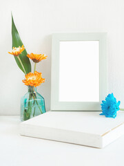 White wooden rectangle photo frame and orange flower, blue (chrysanthemum) with green leaves in vase on a wooden shelf with a white wall as background..