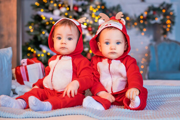 twins toddlers in red santa's reindeer suits sit next to each other at home against the background of christmas tree