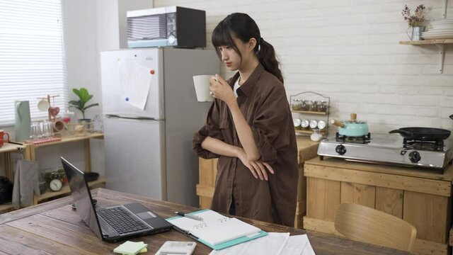 Asian woman creative worker sipping tea and looking at computer in contemplation with an arm on chest is bending over to type on keyboard while working from home