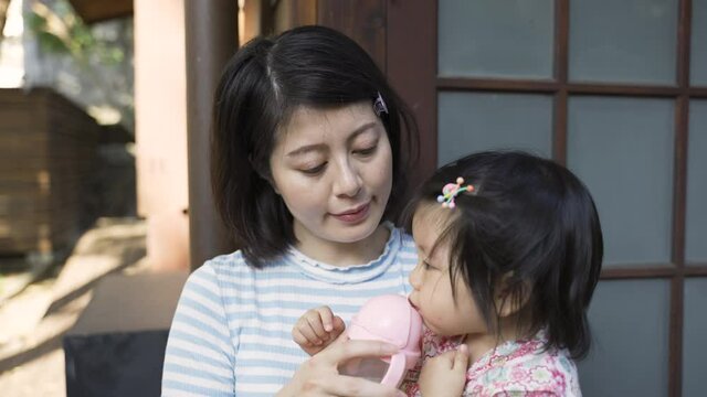 closeup of a japanese mom giving her thirsty toddler baby straw sipper cup with water while taking break at their japanese style house on a sunny day in summer