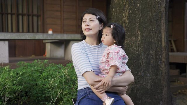happy japanese mother with her toddler kid in arms is looking into distance while sitting under the tree to enjoy breeze in a nearby neighborhood park.