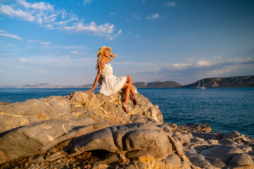 Beautiful young woman wearing traw hat sitting in rocks in white dress by the seashore