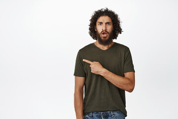 Shocked and worried middle-eastern guy pointing finger left, looking upset and confused, standing over white background