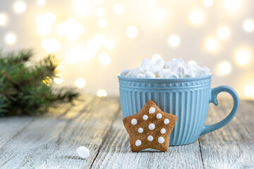 A blue cup of winter hot drink with marshmallows and gingerbread star on a white wooden background with bokeh.