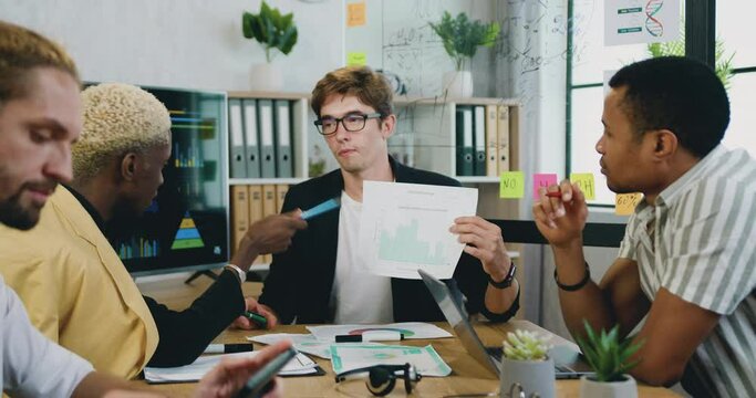 Good-looking confident qualified young male office manager discussing financial report with charts and datas on interactive monitor together with his multiethnic male colleagues in office