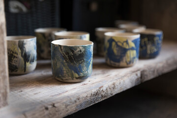 Handmade empty clay mugs composition, painted with blue, green and yellow, on a wooden shelf in a shop