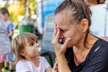 A frustrated young mother struggles with life's problems while spending time with her children outside, the youngest daughter gives her support by hugging her. Motherhood concept.