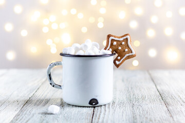Obraz na płótnie Canvas A white cup of winter hot drink with marshmallows and gingerbread star on a white wooden background with bokeh.